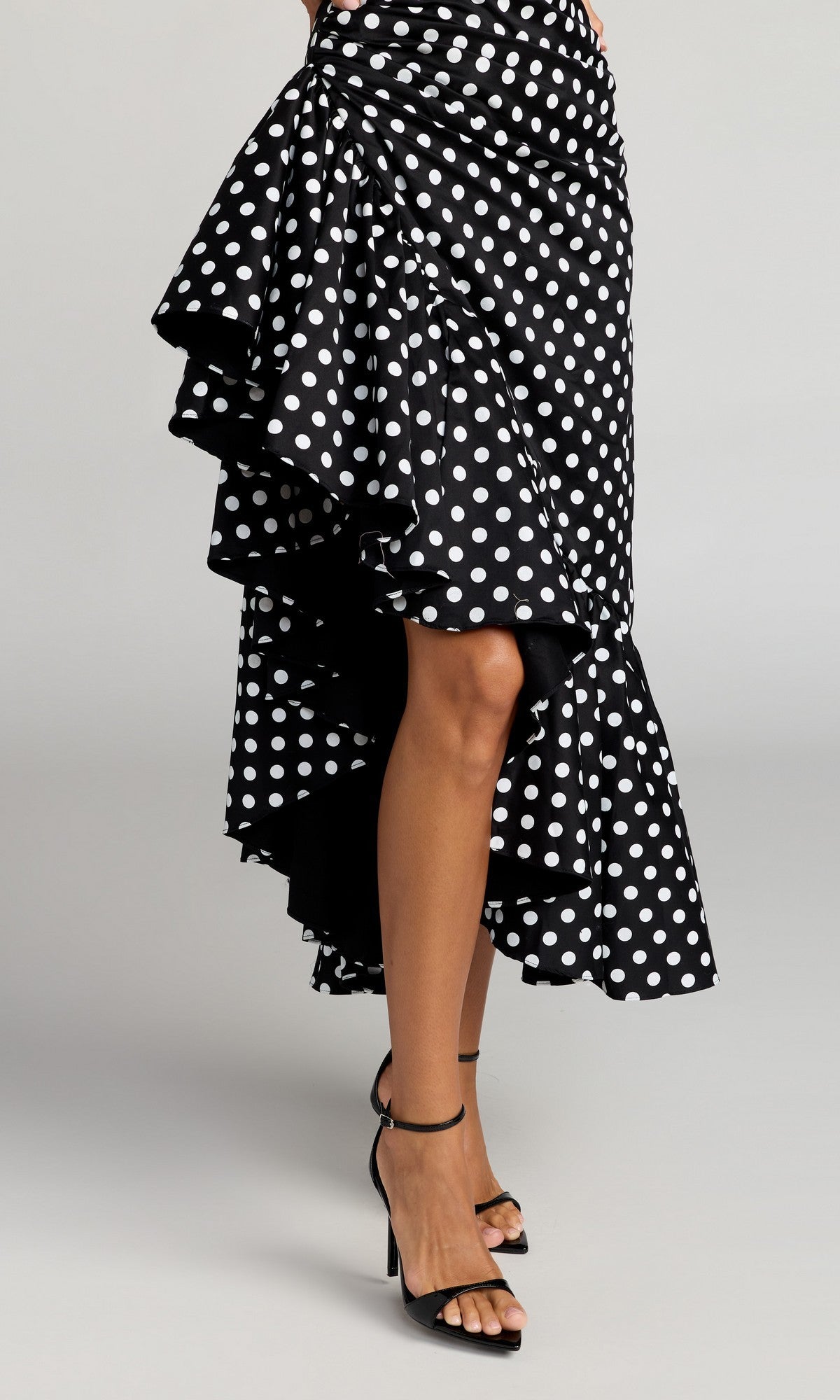 A person wearing the Mac Duggal 27780 Midi Party Dress—a black cocktail dress with a high-low ruffled skirt adorned with white polka dots—paired with black high-heeled sandals, photographed against a plain background.